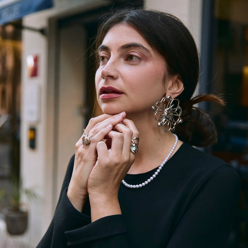 Woman wearing flower earring, 2 rings and a pearl necklace, sitting outdoors with a blurred background
